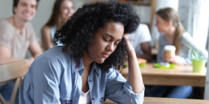 A young woman sits with her eyes closed and her left hand propped against her head. In the background, four people talk with each other.