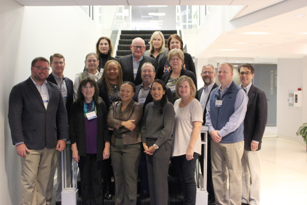 A group of 17 people are gathered in front of and around a flight of stairs and smiling.